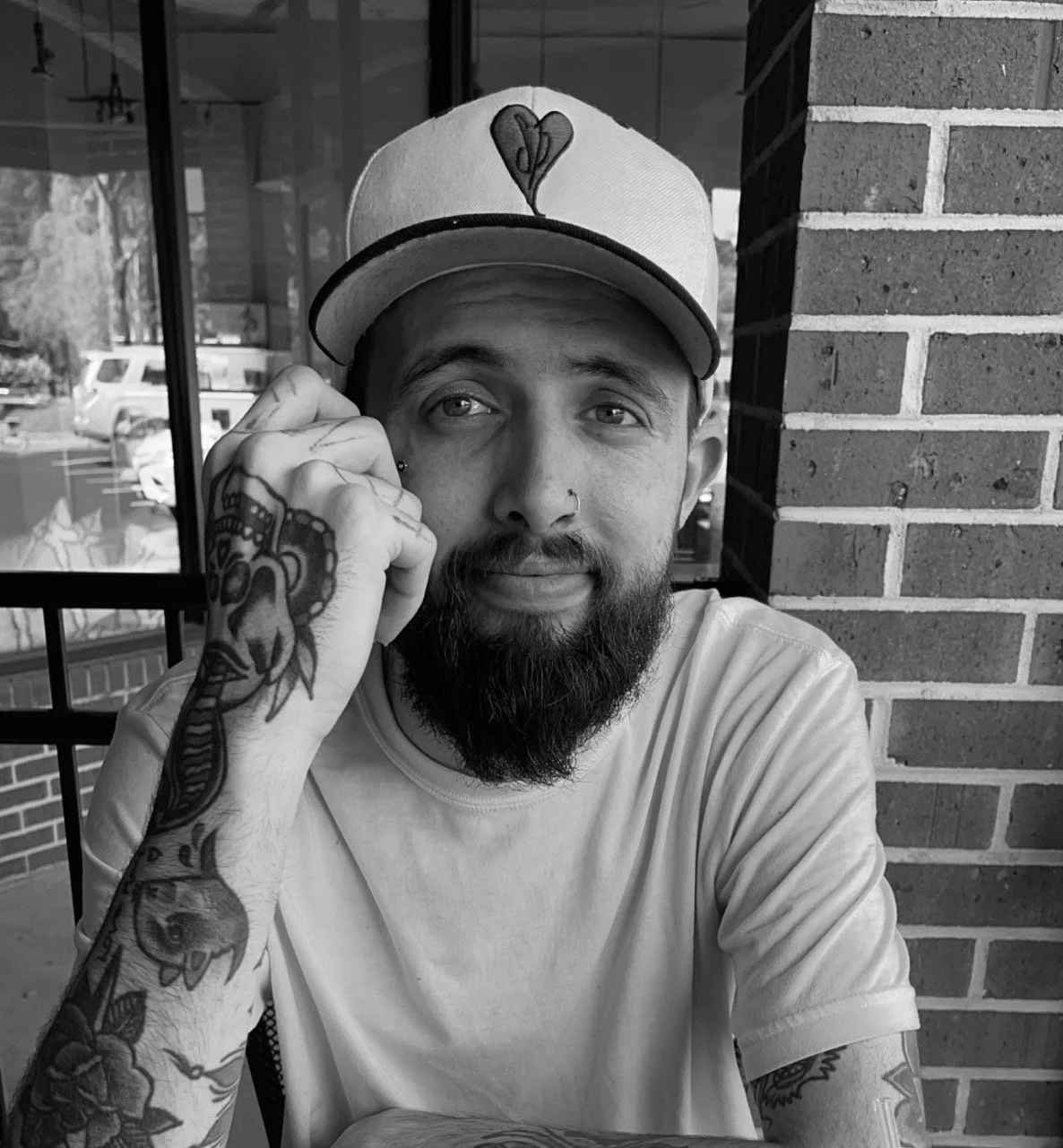 A bearded man wearing a cap with a heart logo, sitting at an outdoor cafe table, resting his head on his hand, with tattoos on his arm and a nose piercing.