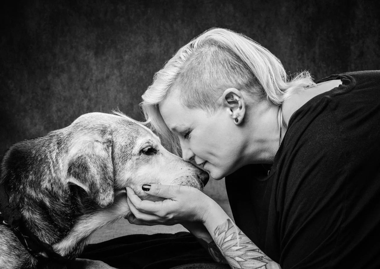 A woman with short, light-colored hair and tattoos gently touching her dog's face with her hands, touching foreheads in a close moment.