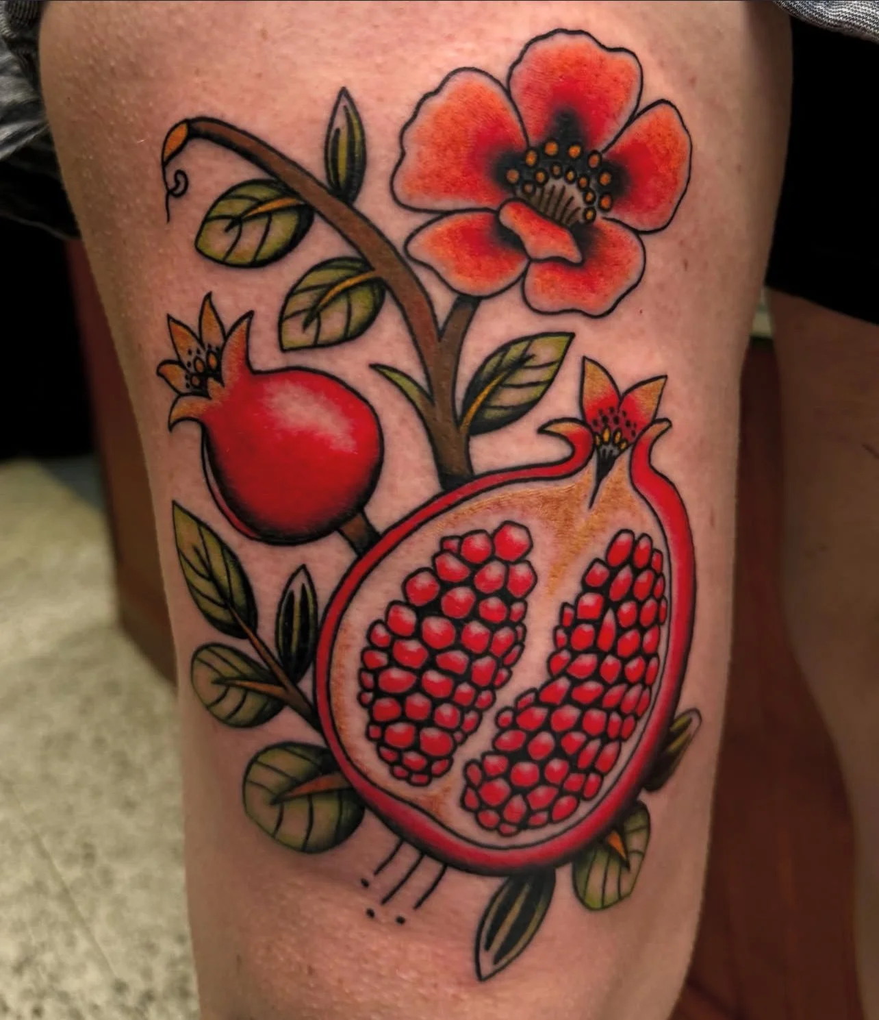 Colorful tattoo of a pomegranate, its seeds exposed, along with pomegranate flowers and leaves.