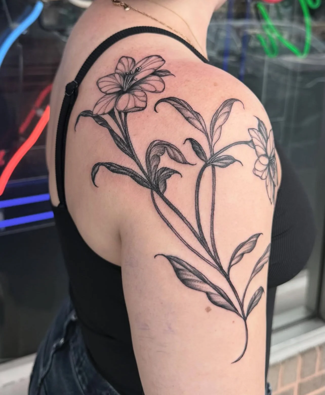 Black ink floral tattoo of two large flowers with multiple leaves on the upper arm of a person wearing a black tank top.