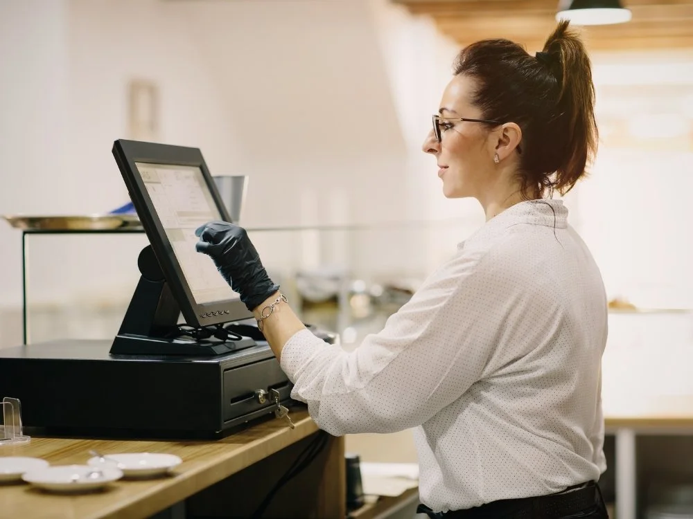 Cashier at a point of sale terminal with a touchscreen interface, wearing a white shirt and black gloves, standing behind a counter in a retail or restaurant setting.