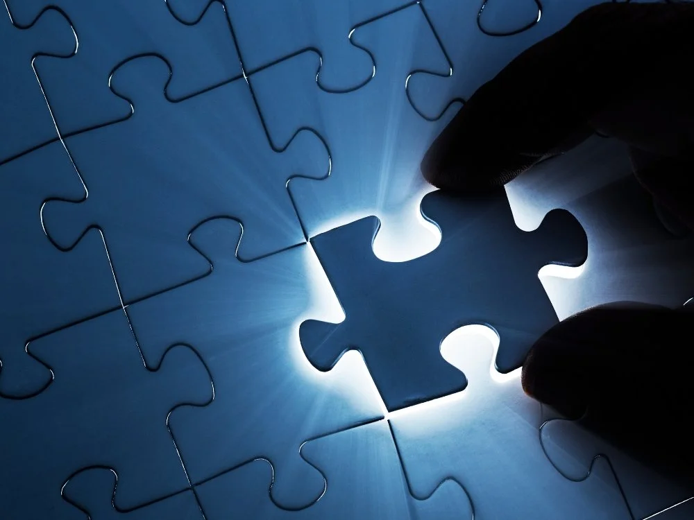 A hand fitting the last puzzle piece illuminated with light shining through, completing a blue puzzle.