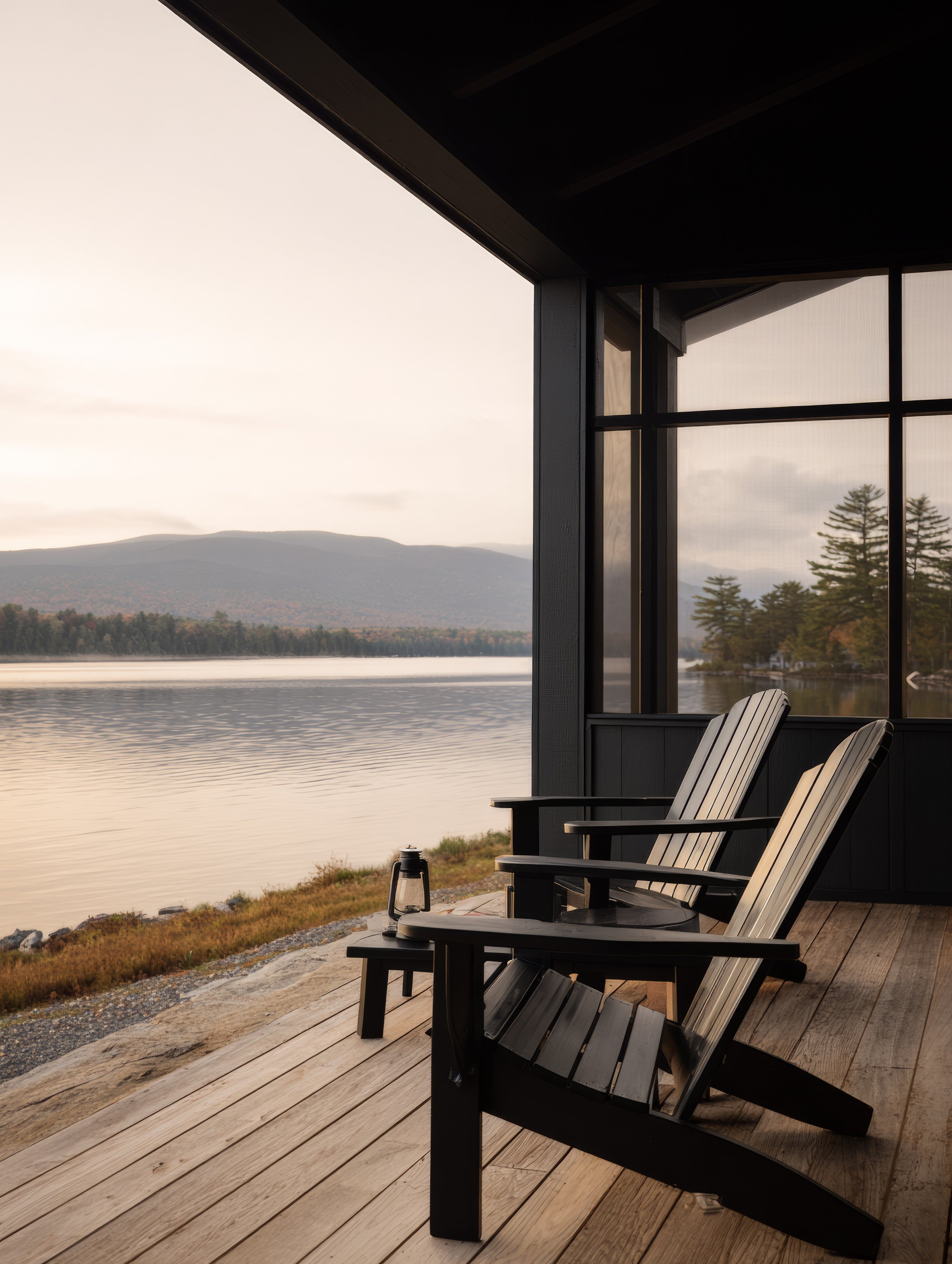 Two Adirondack chairs on a wooden deck overlooking a lake with mountains in the distance, during sunset, with trees reflected in the water.