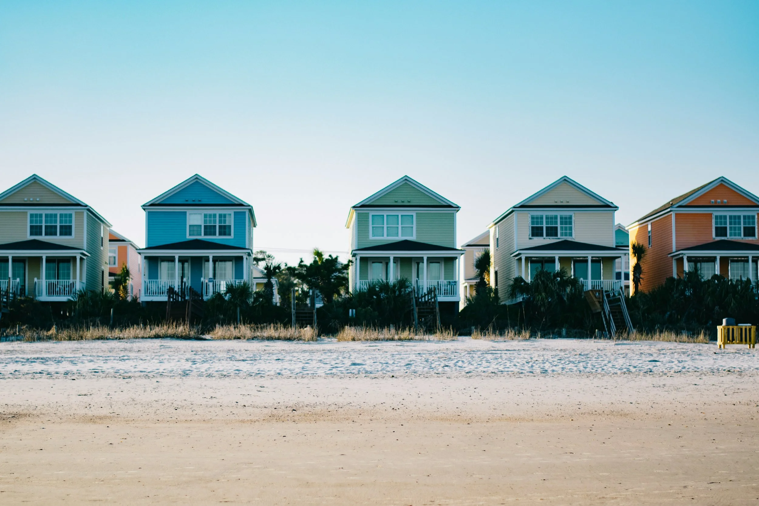 Colorful beach houses on stilts behind a sandy beach, with blue sky overhead.