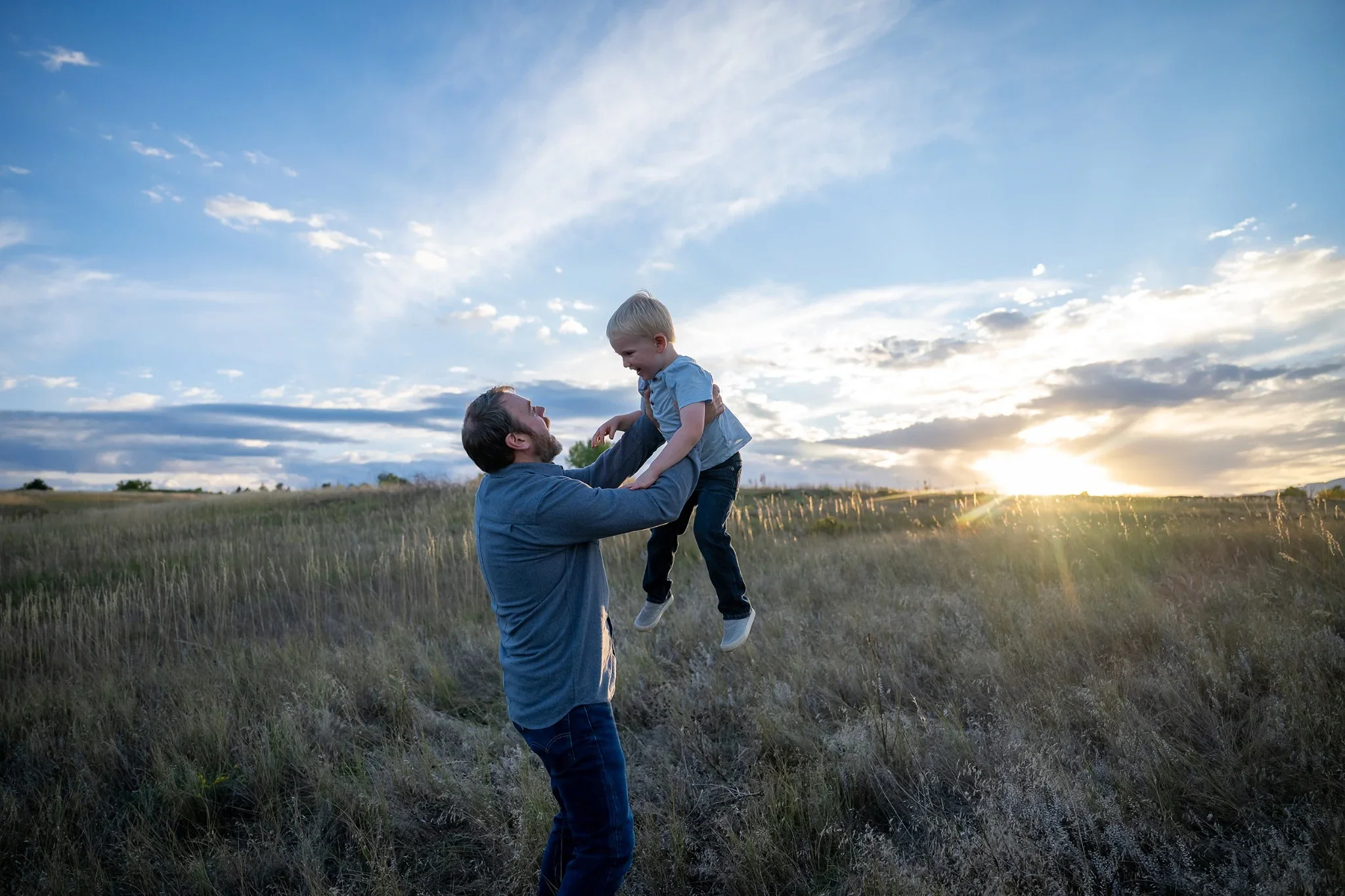 Dad lifts sun up high off the ground as the setting sun dips below the horizon at Standley Lake in Westminster, Colorado Relaxed, natural family photography captured by Ryan Kost.
