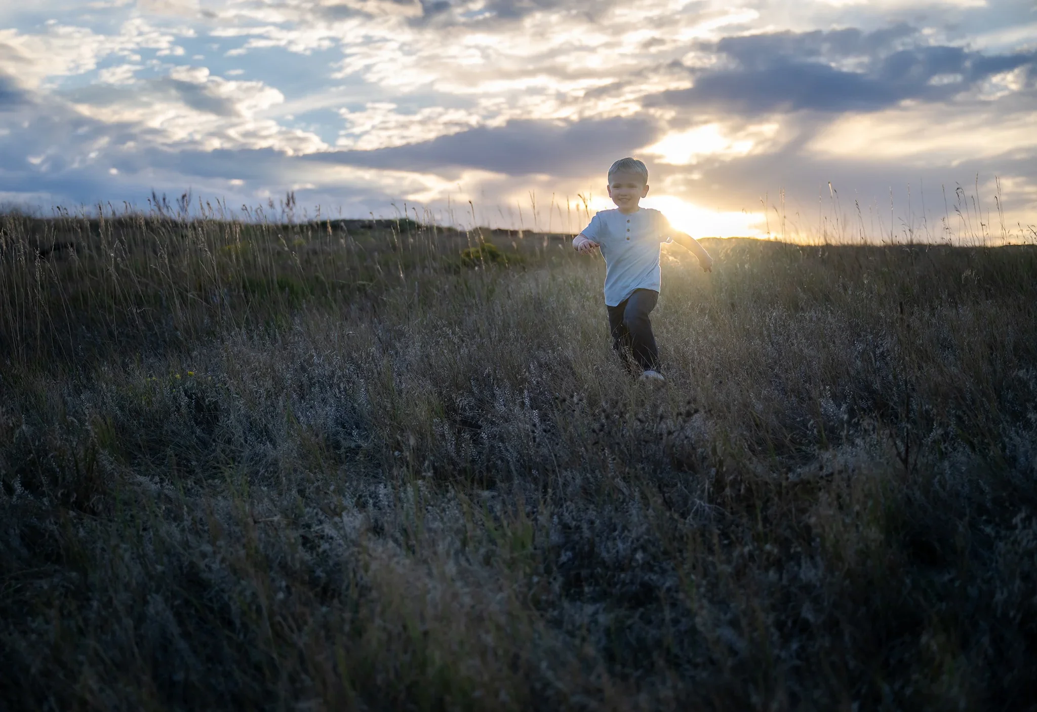 Boy running in field with setting sun behind him at Standley Lake in Westminster, Colorado. Relaxed, natural family photography captured by Ryan Kost
