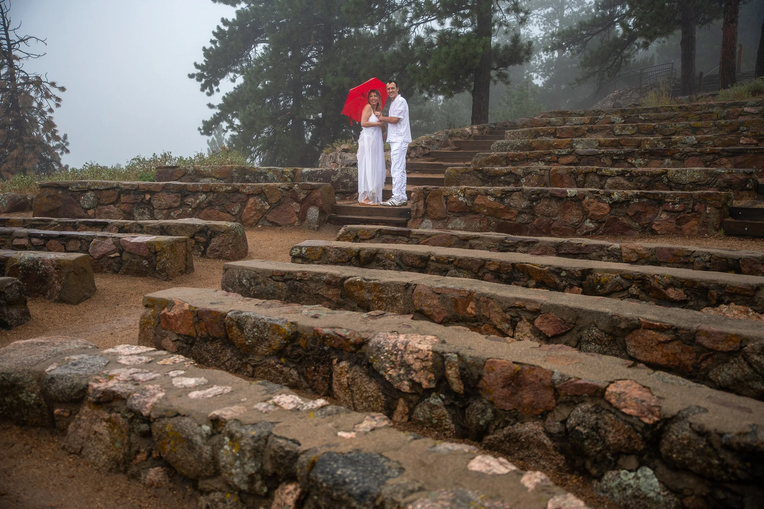A couple dressed in white standing on stone steps in a foggy outdoor setting, holding a red umbrella.