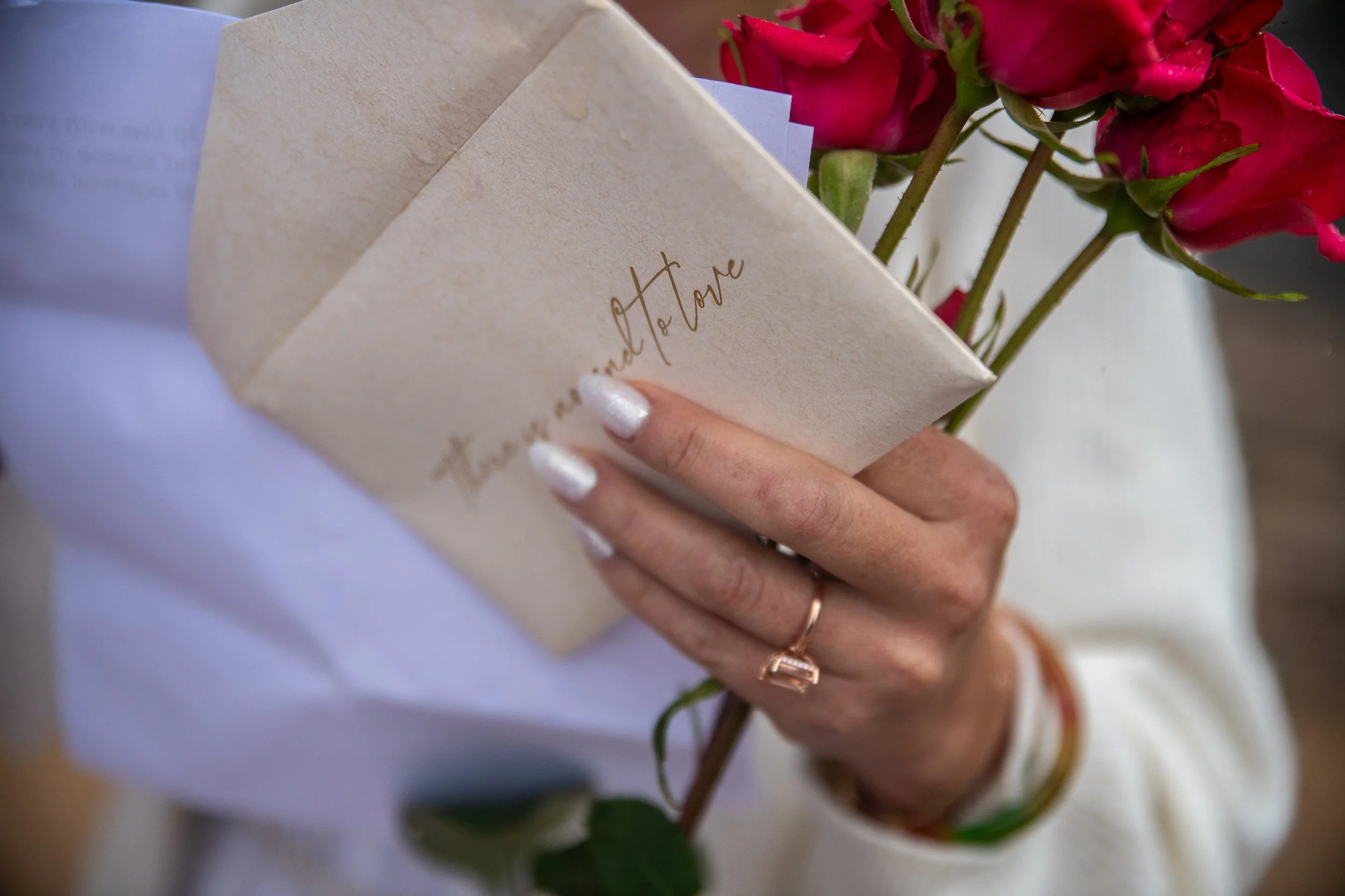 Close-up of a person's hand holding a cream-colored envelope with the words 'trust and love' written on it, a bouquet of pink roses, and the person is wearing rings and nail polish.