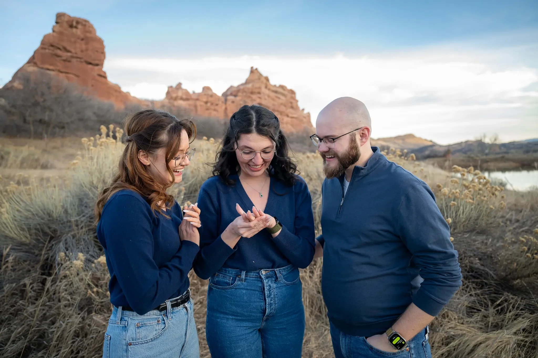 A couple looking at engagement ring with sister-in-law moments after surprise proposal at South Valley Park in Littleton, Colorado captured by Ryan Kost.