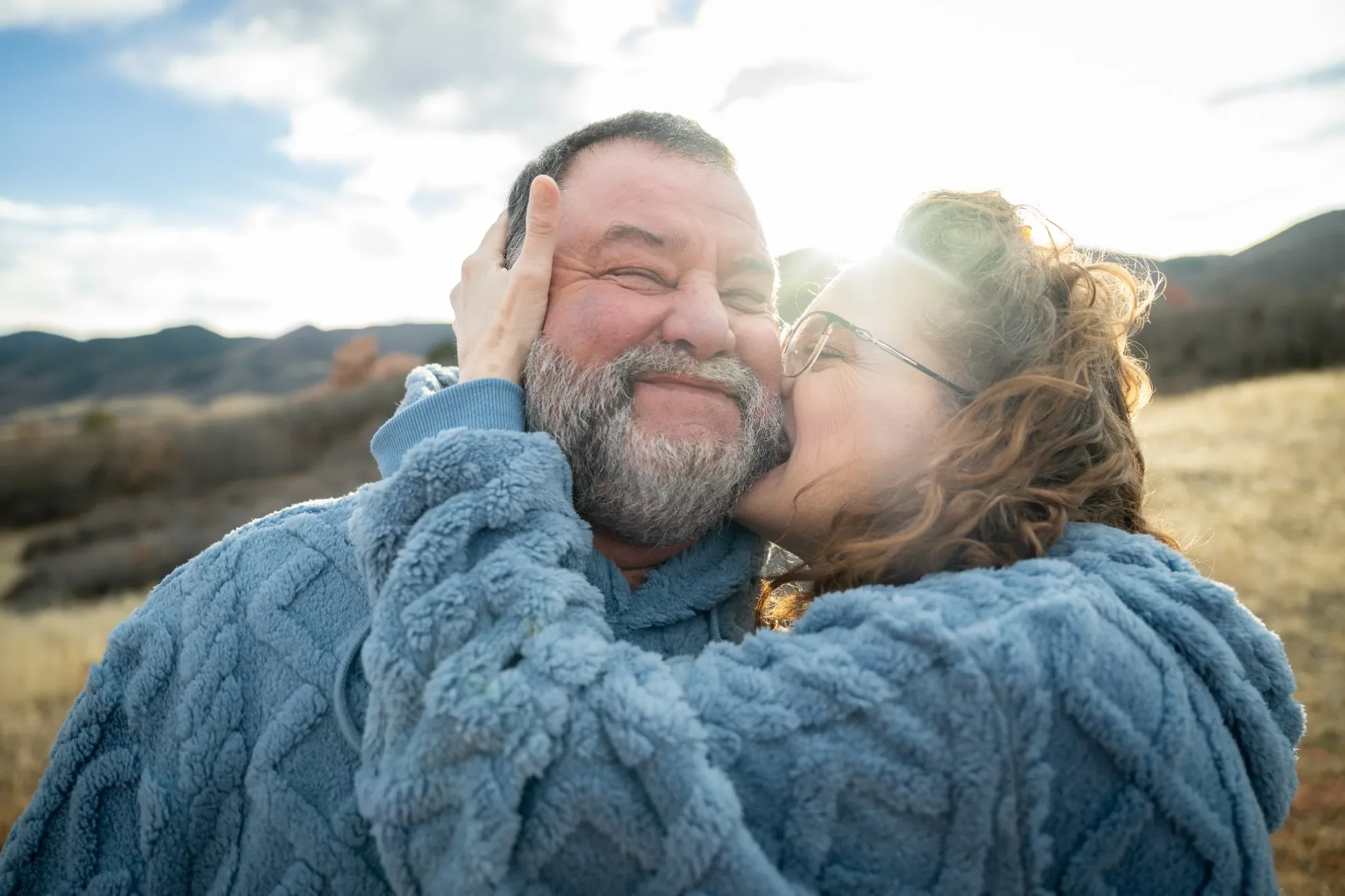 A couple hugging outdoors in a field with mountains in the background, enjoying a happy moment together.