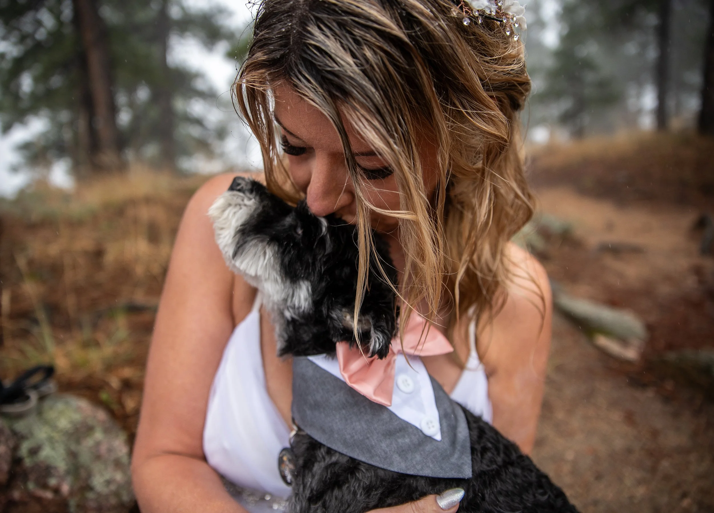 A woman holding a small black and white dog, both with their eyes closed, in an outdoor wooded area.