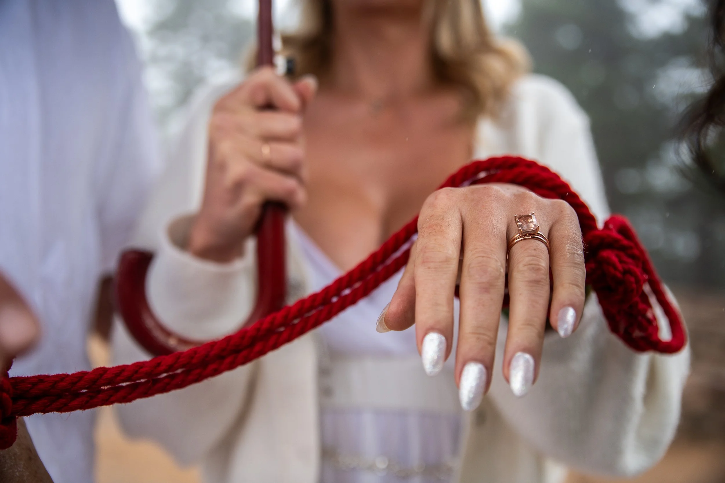 A woman's hand with a large gemstone ring on her ring finger, holding a thick red rope. She is dressed in white, and the background is blurred with trees.
