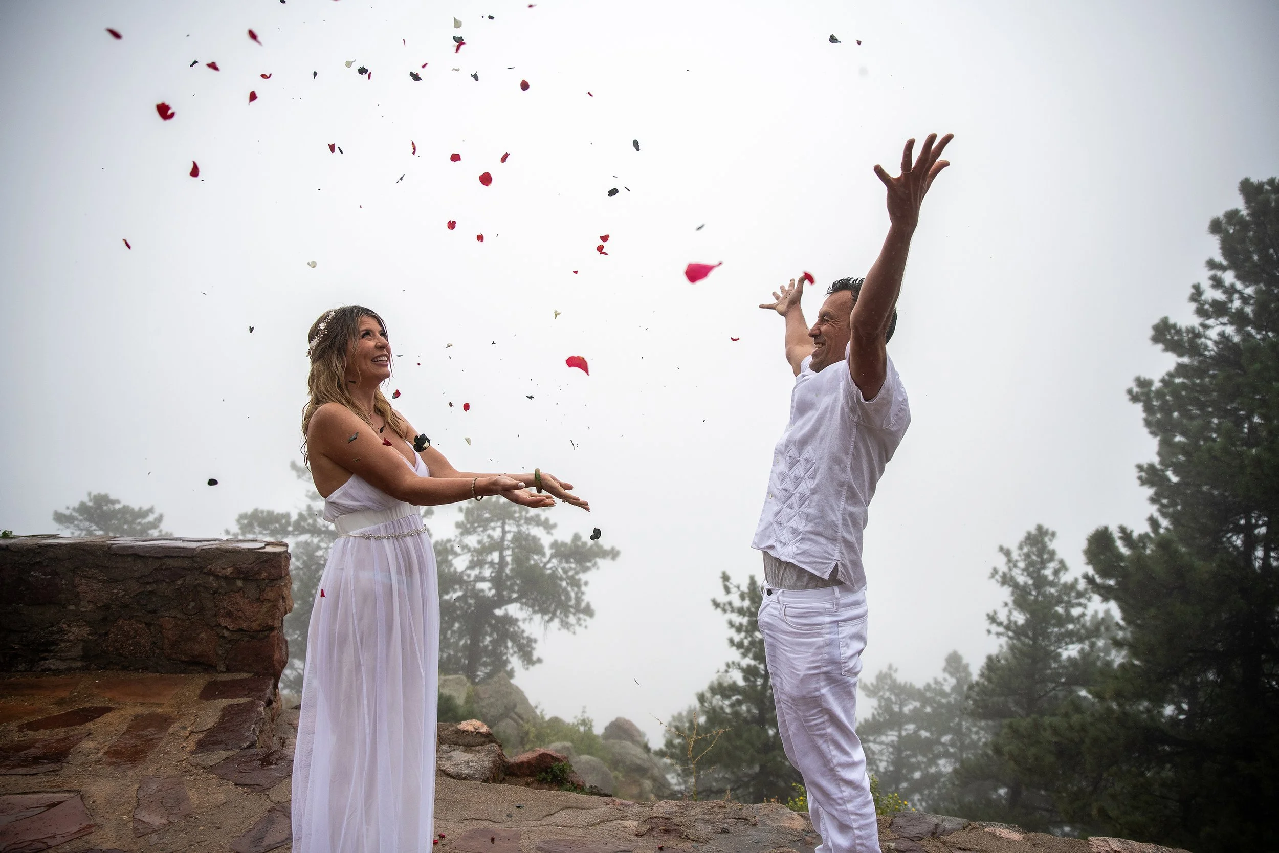 A man and woman in white celebrating outdoors with rose petals in the air amidst a foggy forest landscape.