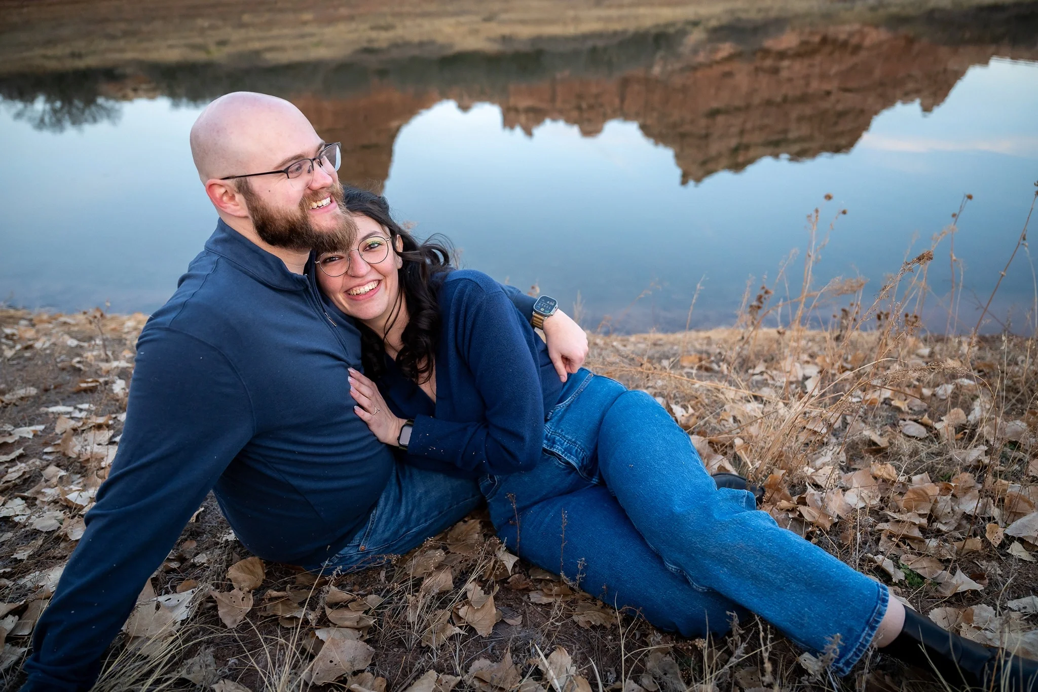 Engaged couple embracing beside a quiet lake at South Valley Park in Littleton, Colorado, photographed in a natural documentary style by Ryan Kost