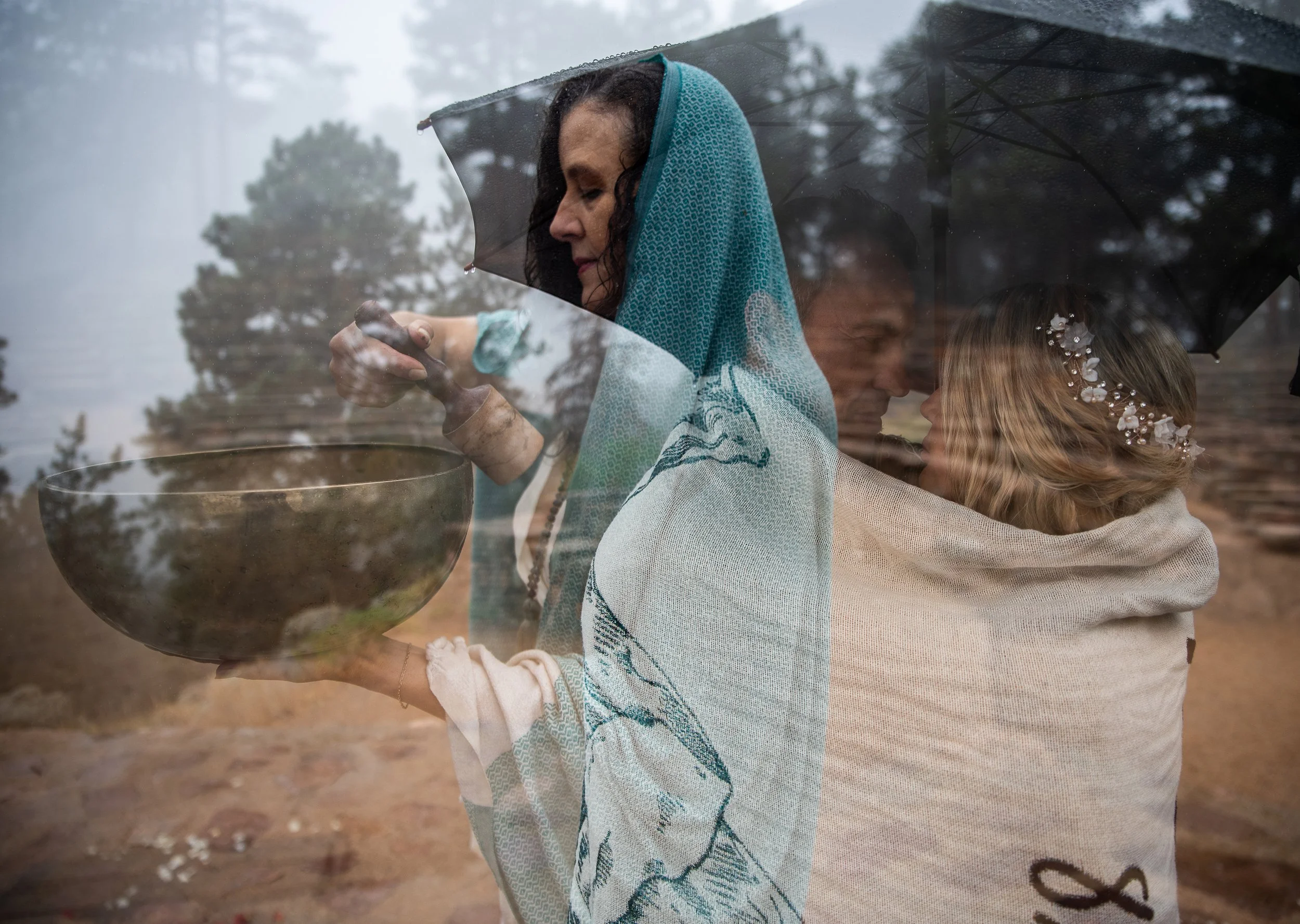 A woman in a blue shawl holding a bowl, with a priest or officiant and a girl in ceremonial attire under an umbrella outdoors.