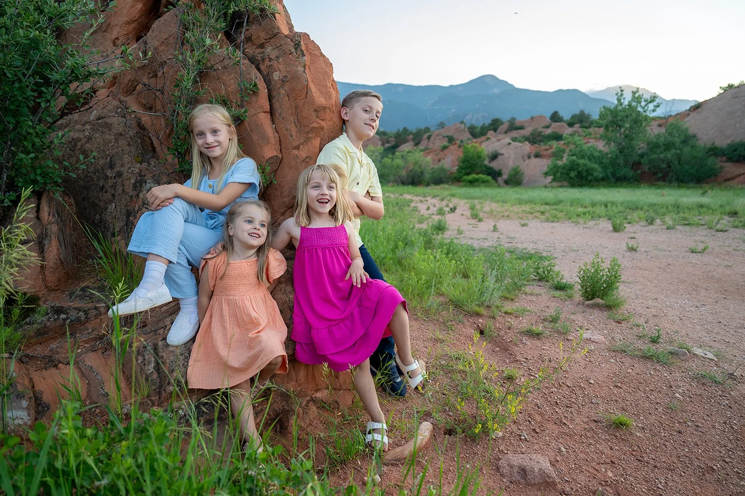 Colorful siblings leaning against red rock formations during a summer family photo session at Red Rock Canyon Open Space in Colorado Springs, with foothills in the background. Captured by Ryan Kost Photography”