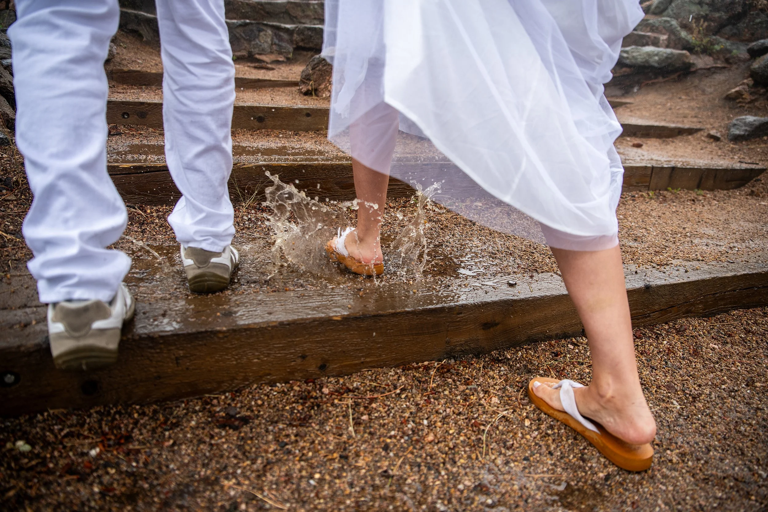 People wearing white clothes and sandals walking through water on a muddy outdoor staircase.