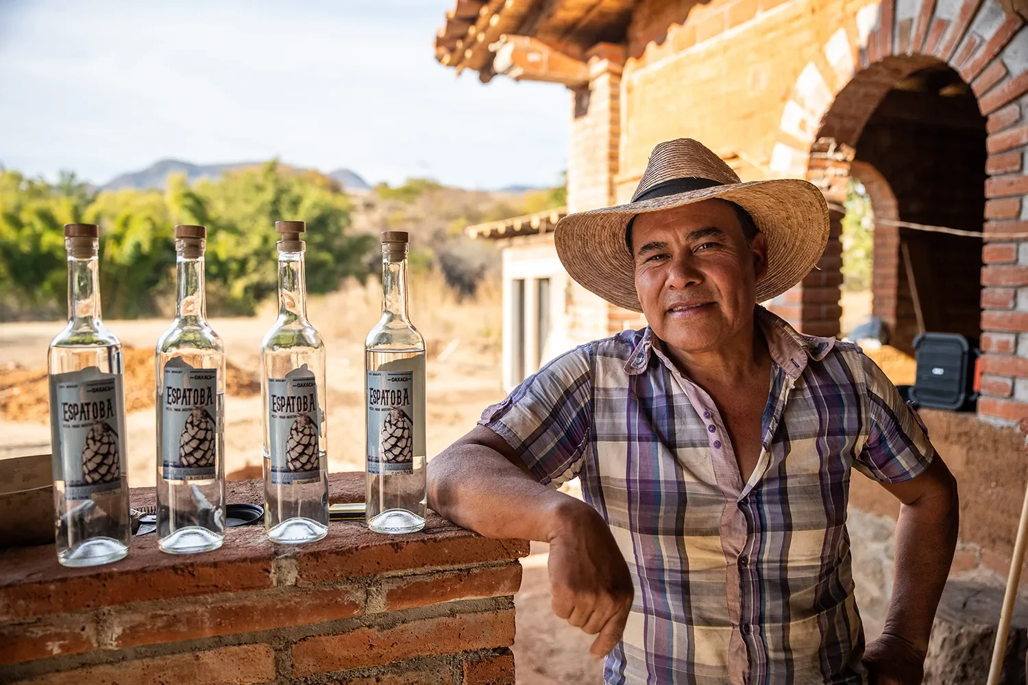 A man wearing a straw hat and a plaid shirt standing outdoors next to a brick structure, with four bottles of clear liquid on a brick ledge in front of him, in a rural setting with trees and hills in the background.