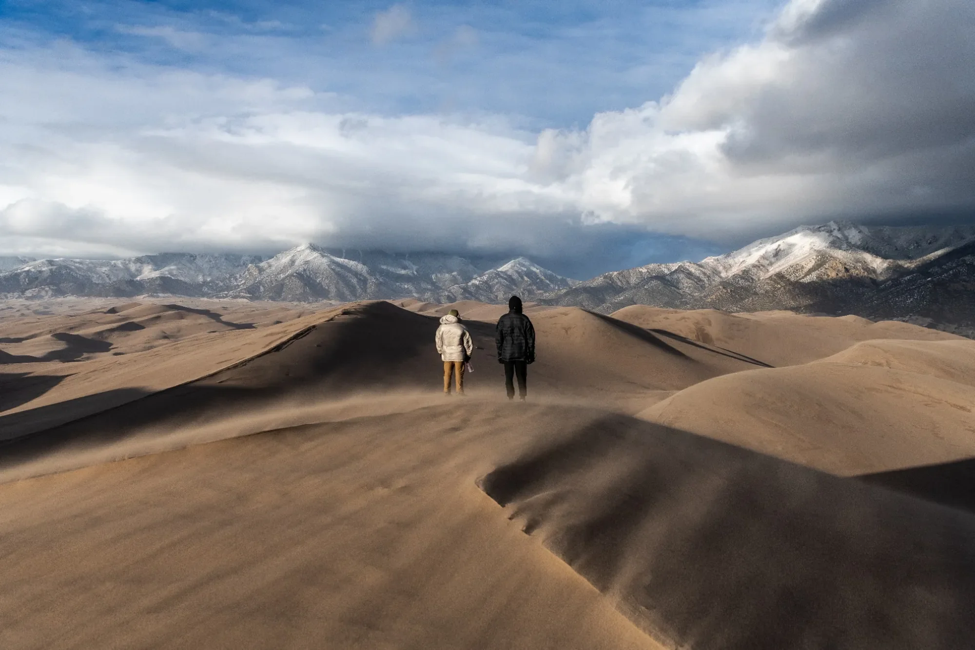 Father and son visiting Great Sand Dunes National Park in Colorado after traveling from Peru, standing together on windswept sand dunes with mountains behind them