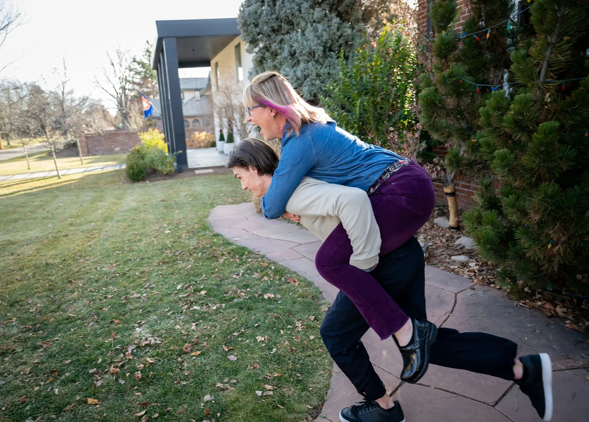 Son running with mom on back. Fun, documentary family photography in yard during a in home family photo session captured by Ryan Kost