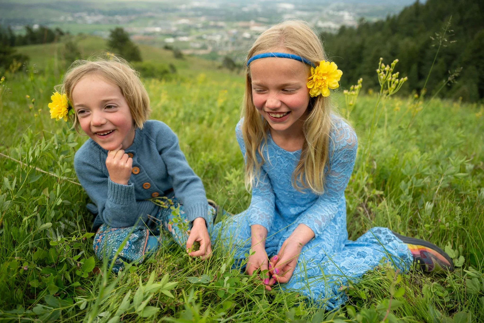 Sisters sitting together in backyard summer meadow picking wildflowers on Lookout Mountain in Golden, Colorado.. Natural, relaxed family photogaphy captured by Denver photographer Ryan Kost.