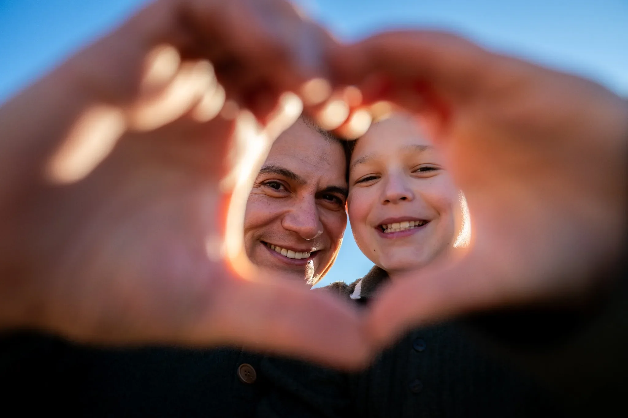 A man and a young boy smiling and looking through their hands, which are forming a heart shape, with clear blue sky in the background.