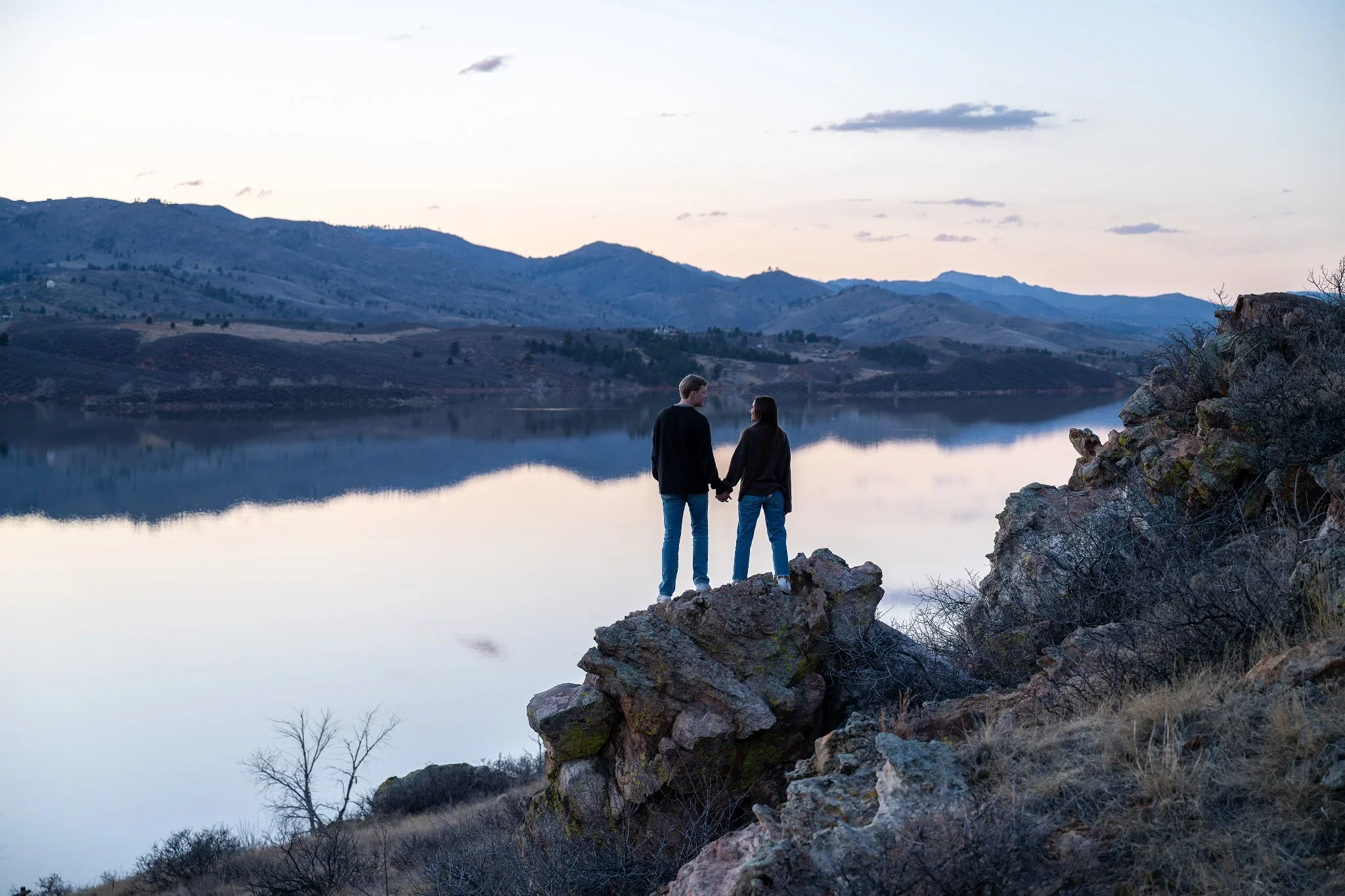 A coule stands on a rock outcropping holding hands taking in the splendor of Horsetooth Reservoir at dusk. Colorado engagement photography captured by Ryan Kost