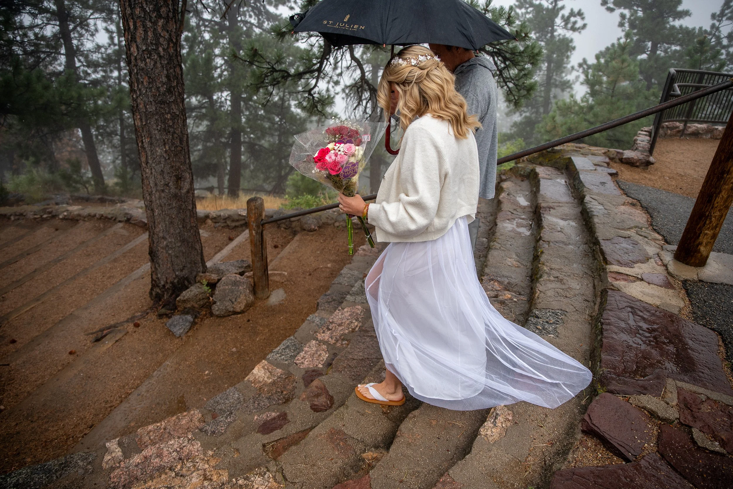 A woman in a white dress, cardigan, and flip-flops holds a bouquet of pink flowers, standing on steps with a man under an umbrella in a foggy, wooded outdoor setting.