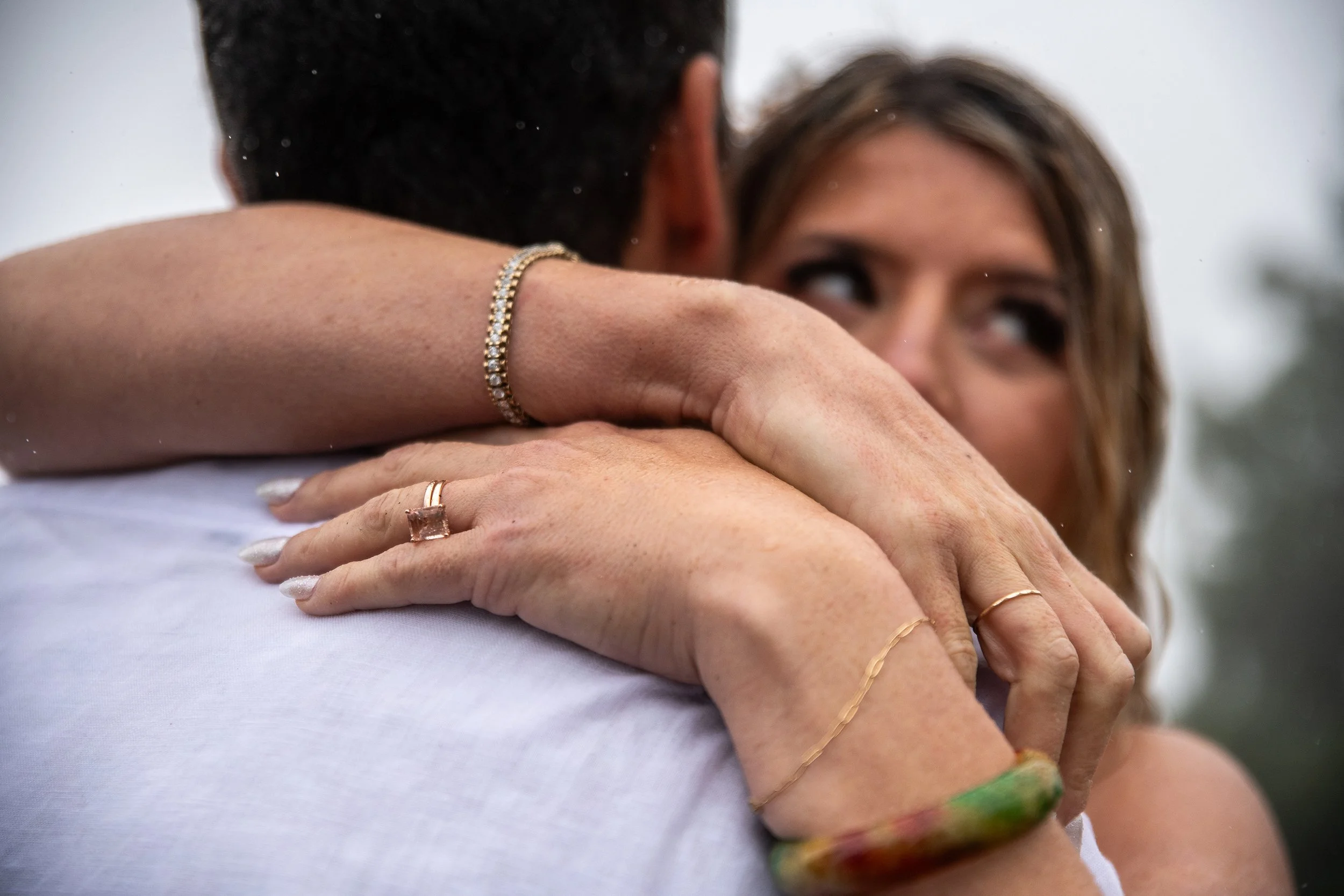 Woman embracing a man with her arms around his shoulders, her face partially visible, wearing jewelry including rings, bracelets, and a necklace.