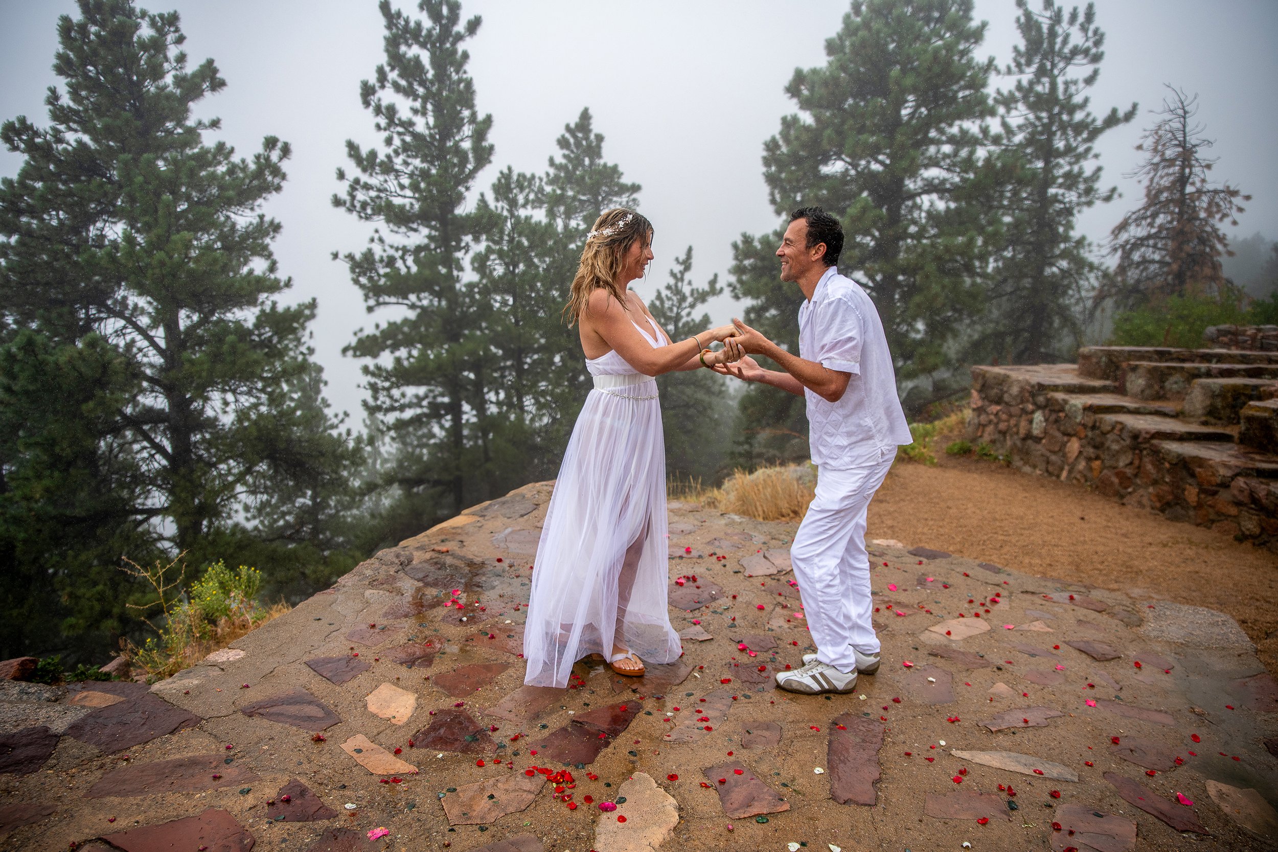A couple in white wedding attire holding hands and smiling on a stone platform outdoors surrounded by pine trees and fog, with rose petals scattered on the ground.