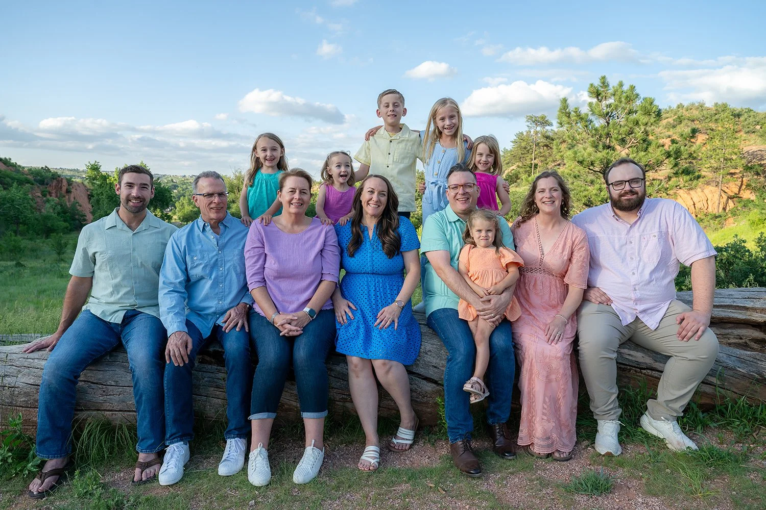 Extended family portrait at Red Rock Canyon Open Space in Colorado Springs with three generations posed on a log, captured in summer evening light by Ryan Kost Photography