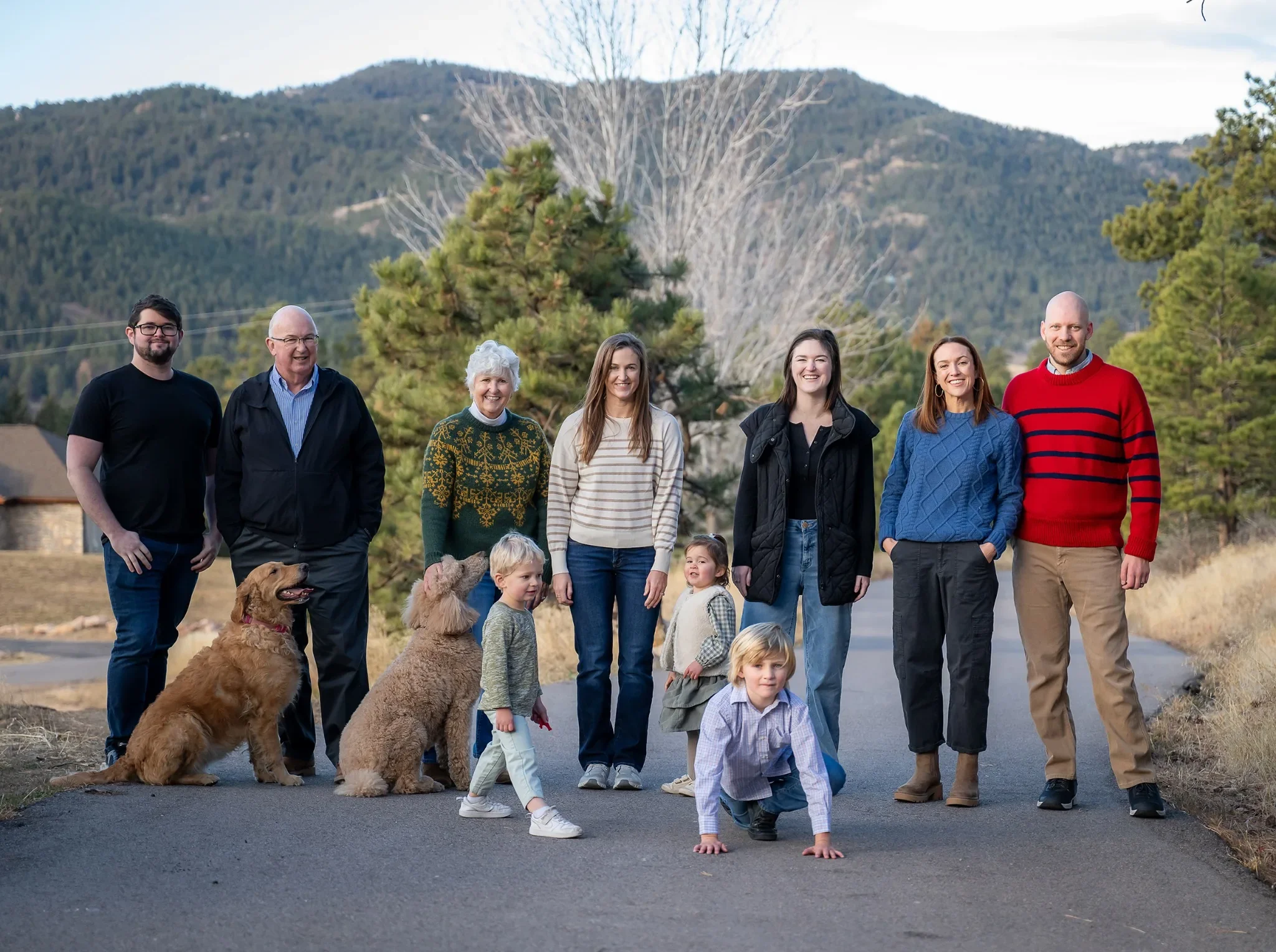 Family photo of eight people with three children and two dogs outdoors on a paved road, with trees and mountains in the background during daytime.