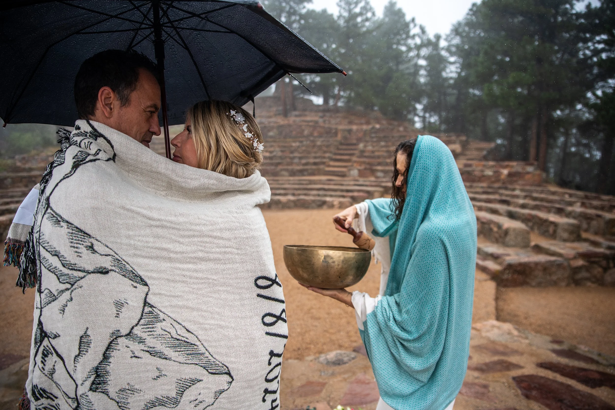A couple is standing outdoors in the rain, sharing an umbrella and wrapped in a white blanket, looking into each other's eyes. A woman dressed in blue is performing a ceremony with a singing bowl in front of them, on a stepped stone structure surroun