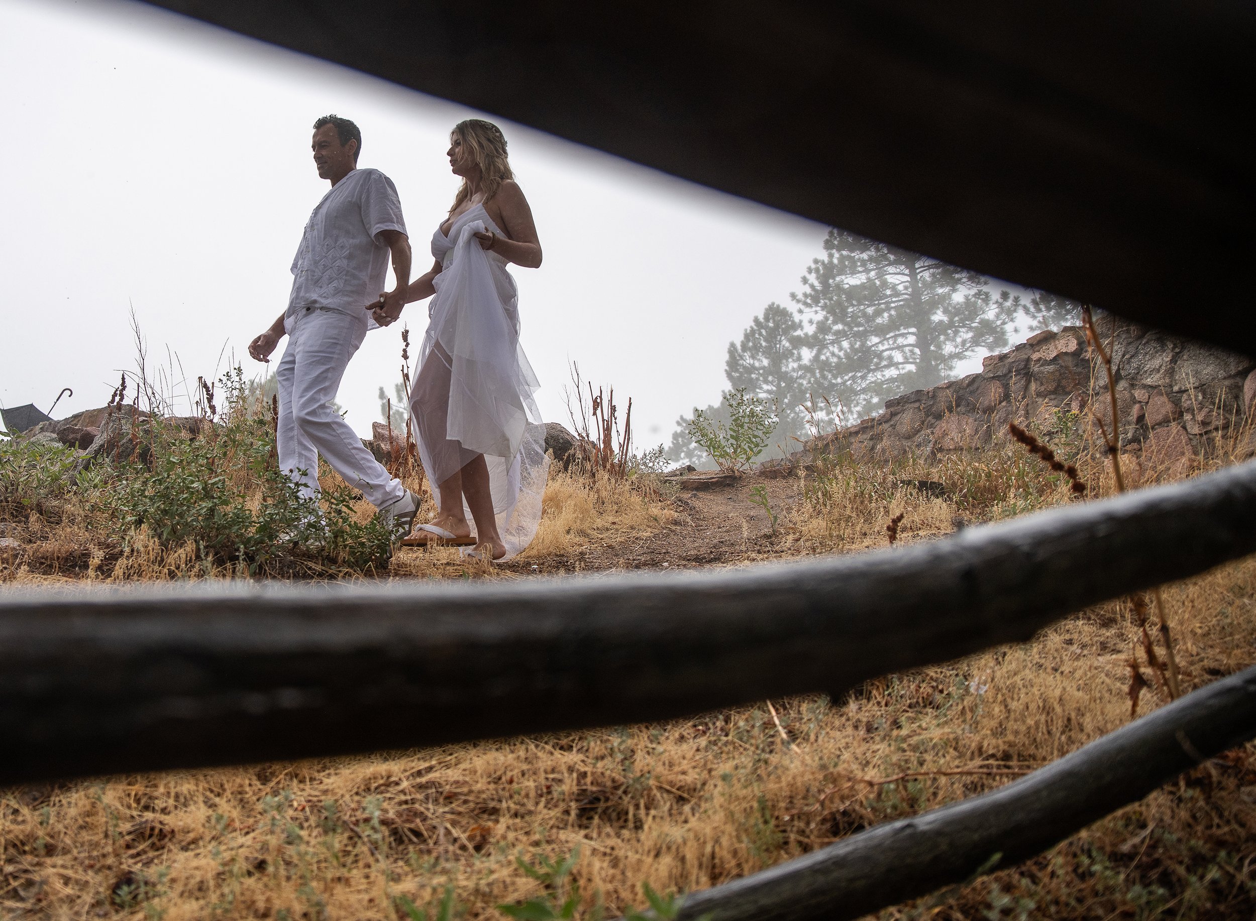 A couple walking hand in hand outdoors on a foggy day, seen through wooden fence rails, with the woman wearing a white dress and the man in white clothing, on a dirt path surrounded by dry grass and rocks.