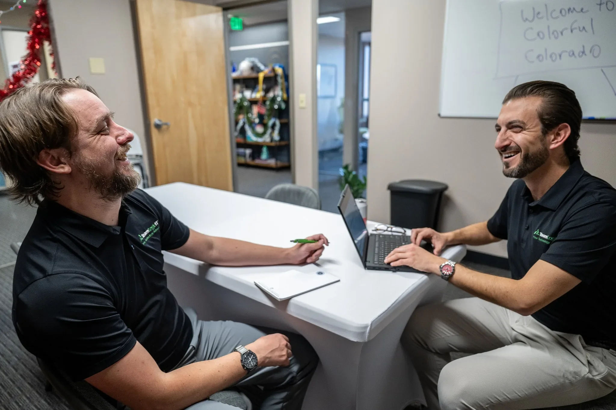 Two men are sitting at a white conference table, laughing and talking. One has a notebook and pen in front of him, the other is using a laptop. The room is decorated with holiday ornaments and a whiteboard in the background reads 'Welcome to Colorful Colorado.'