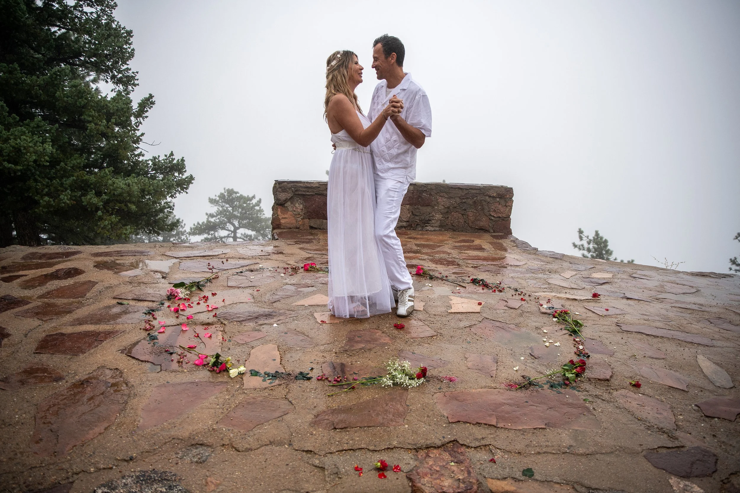 A couple dancing on a stone-paved surface surrounded by scattered flower petals and greenery, with fog and trees in the background.
