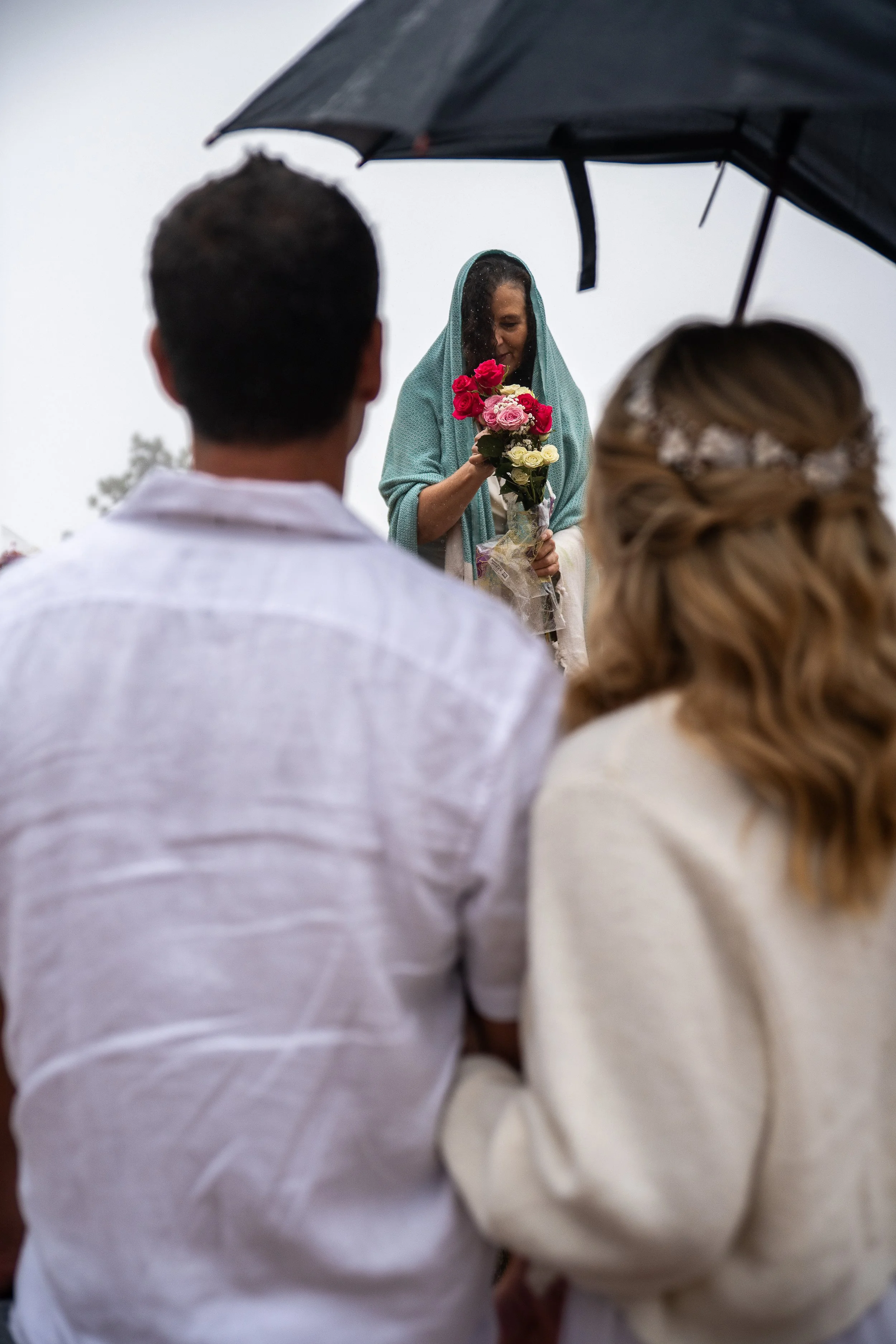 A wedding ceremony on a rainy day with a bride and groom and a woman holding a bouquet of flowers, under an umbrella.