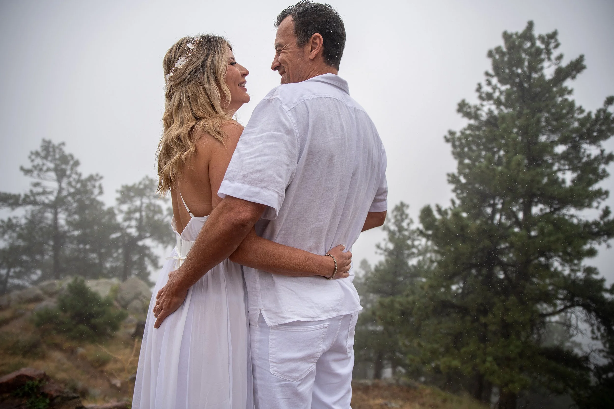 A couple stands close together, smiling and looking at each other, outdoors on a foggy day with trees in the background. The woman wears a white dress with thin straps and a floral headband. The man wears a white shirt and white pants.
