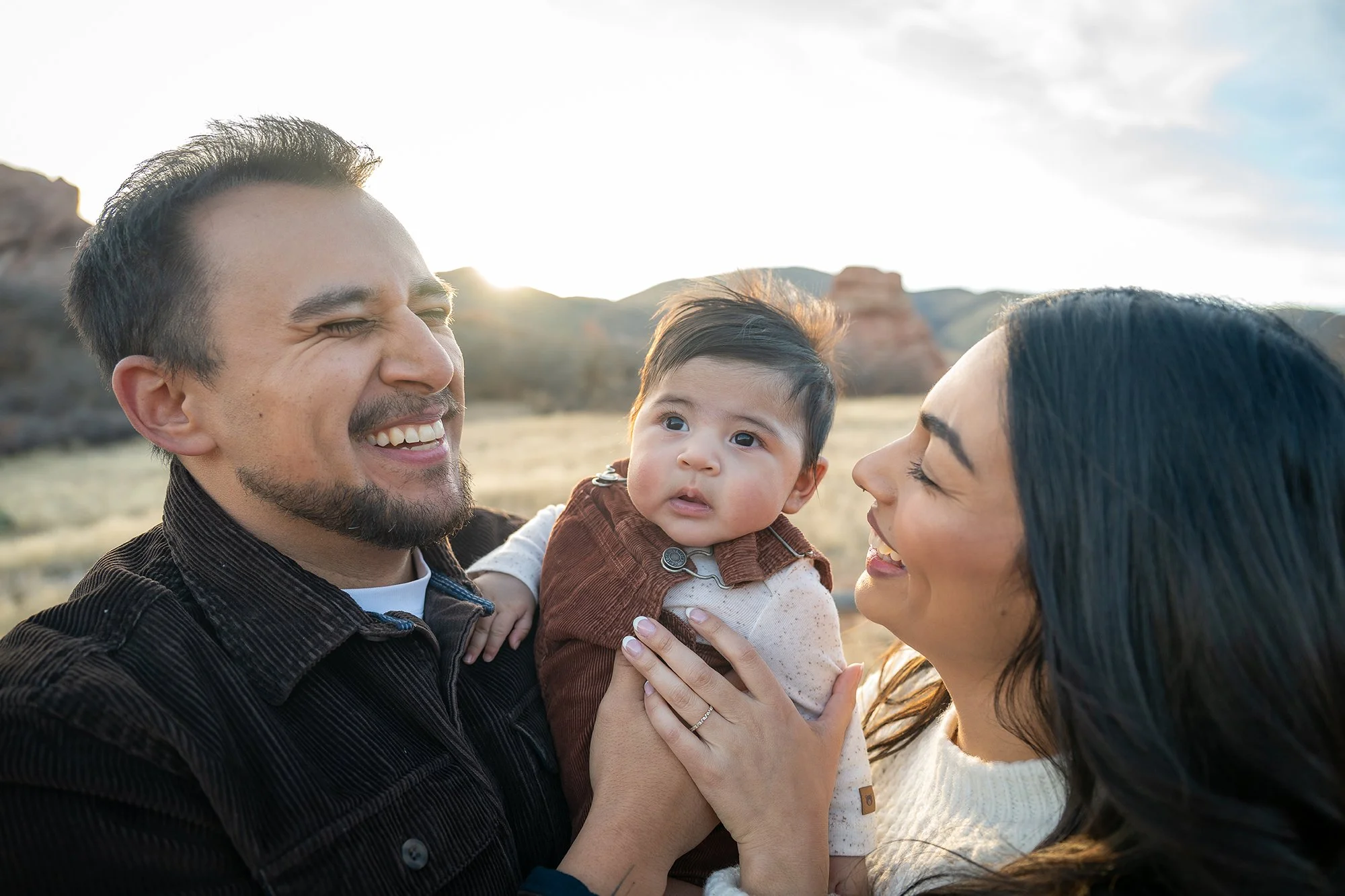 Parents holding infant son sharing loving smiles. Natural, timeless Colorado family portrait photography captured by Ryan Kost at South Valley Park in Littleton.