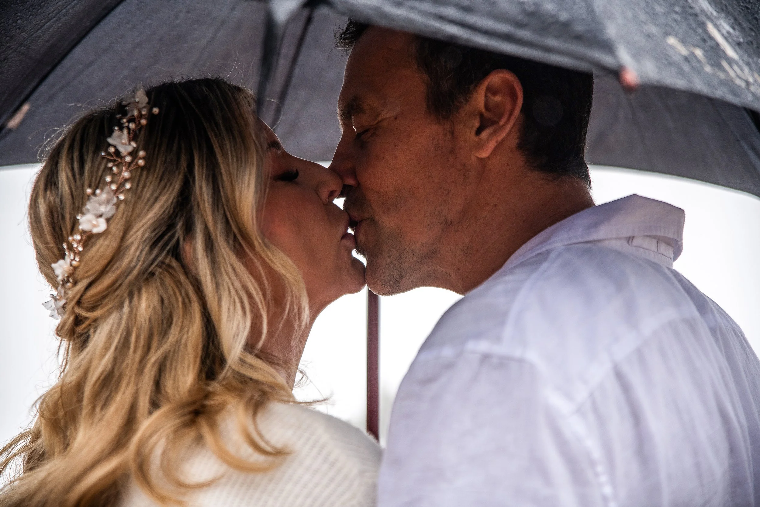 A couple kiss under an umbrella, with the woman wearing a floral headpiece and the man in a white shirt.