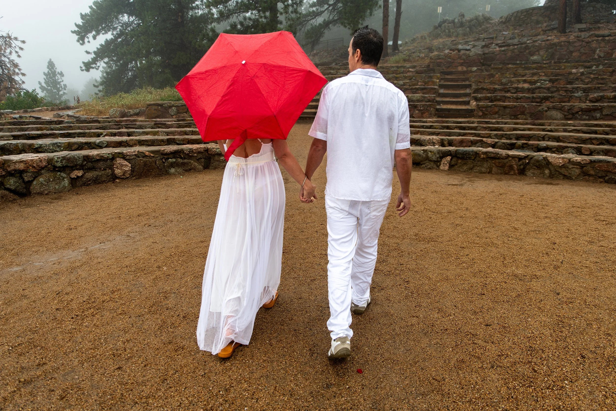 A couple holding hands as it rains, with the woman holding a red umbrella, walking up stone steps outdoors on a misty day.