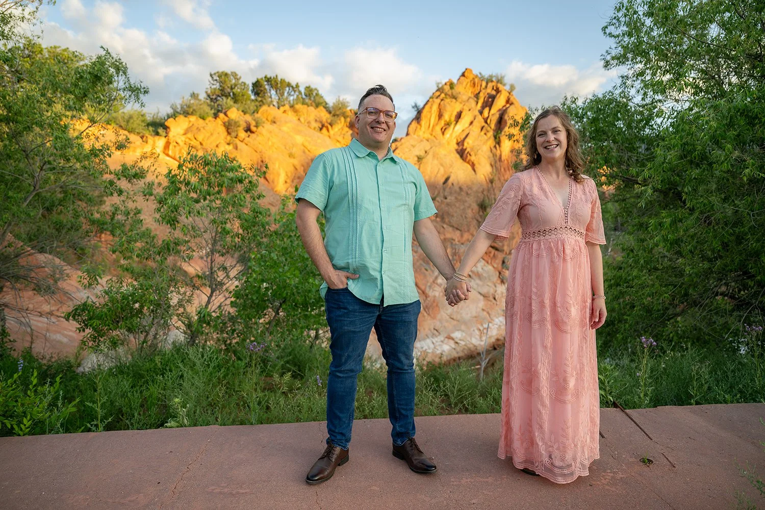Couple holding hands during golden hour at Red Rock Canyon Open Space in Colorado Springs, wearing pastel outfits and smiling in front of scenic red rock cliffs, captured by Ryan Kost Photography