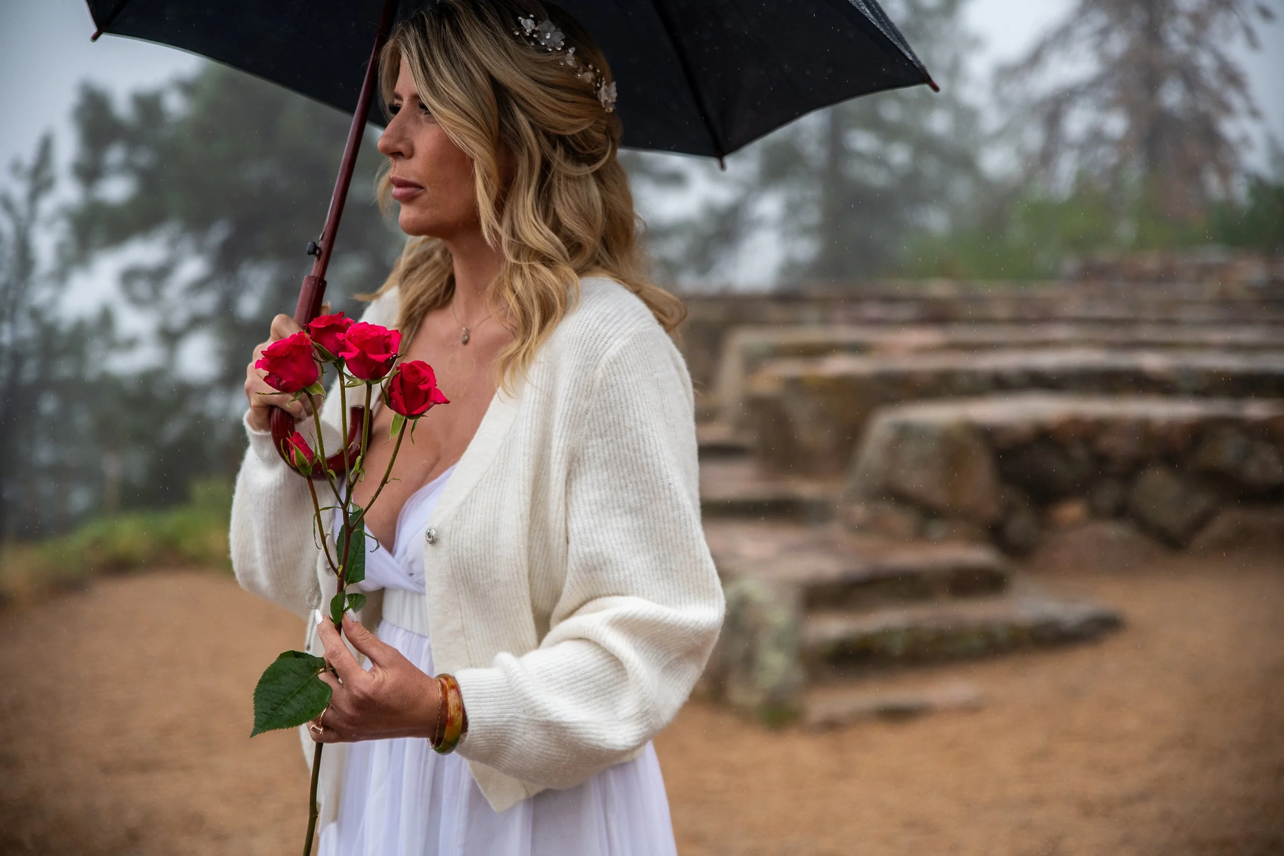 A woman with blonde hair holding a bouquet of pink roses, wearing a white dress and a cream-colored cardigan, stands outside on a rainy day under a black umbrella, with a stone staircase in the background.