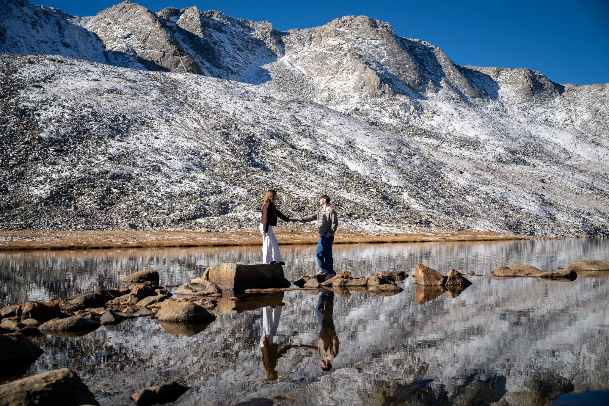 Engaged couple holding hands on rocks at Summit Lake beneath snow-covered peaks on Mount Blue Sky near Denver, Colorado.