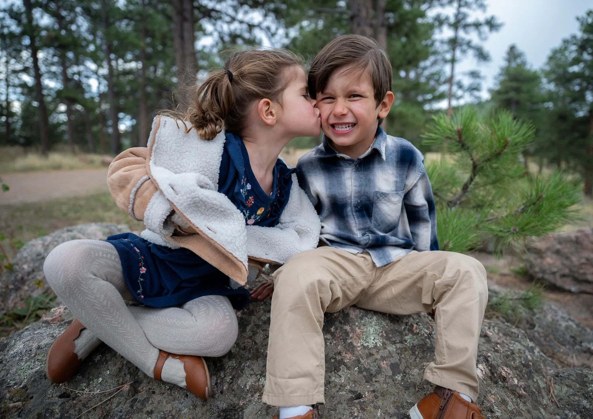 A young girl is kissing a boy on the cheek while sitting on a large rock in a wooded outdoor setting. The boy is smiling with a scrunched face. Both children are dressed in casual, outdoor clothing.