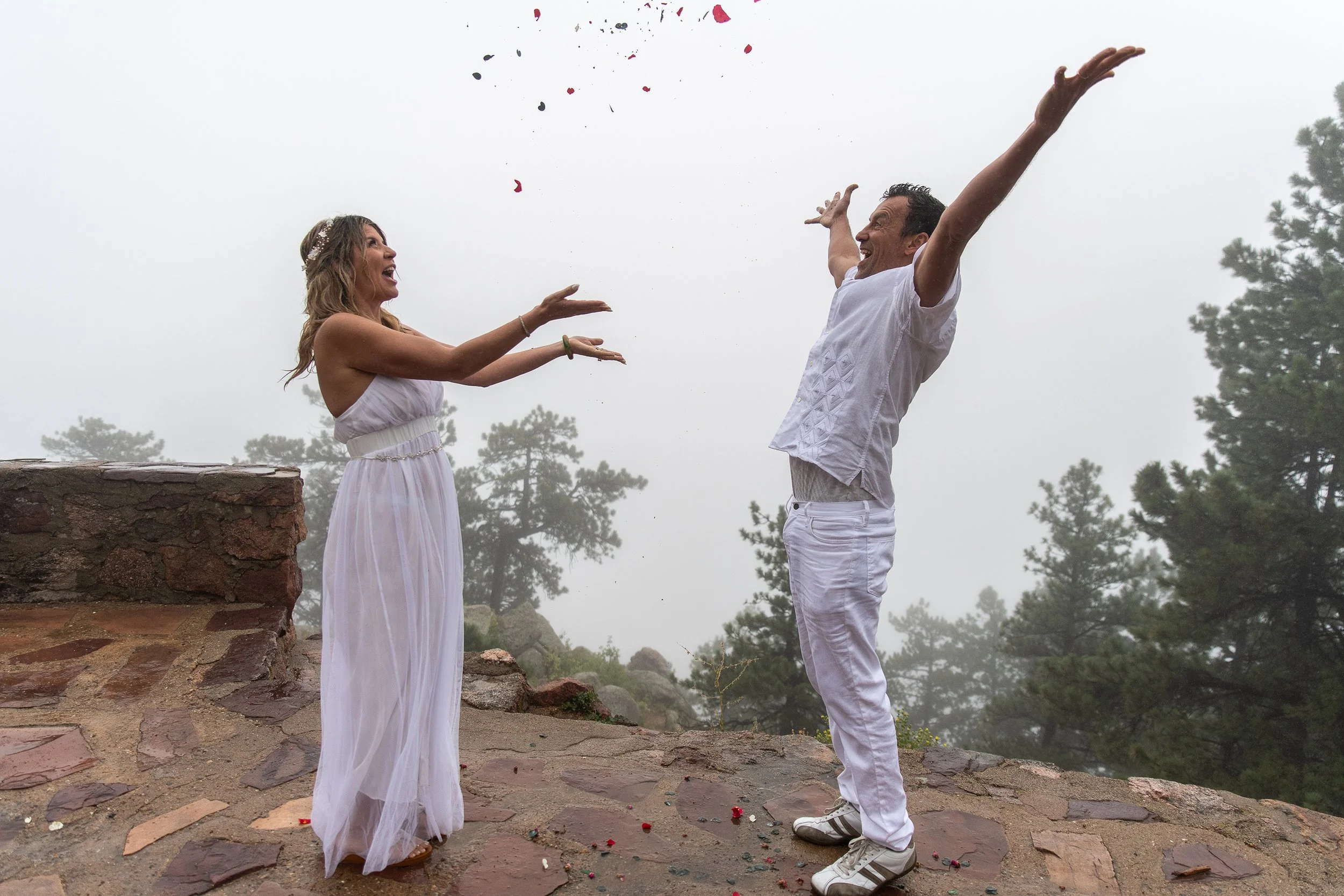A couple celebrating outdoors in a foggy, mountainous area, throwing flower petals into the air with joyful expressions.