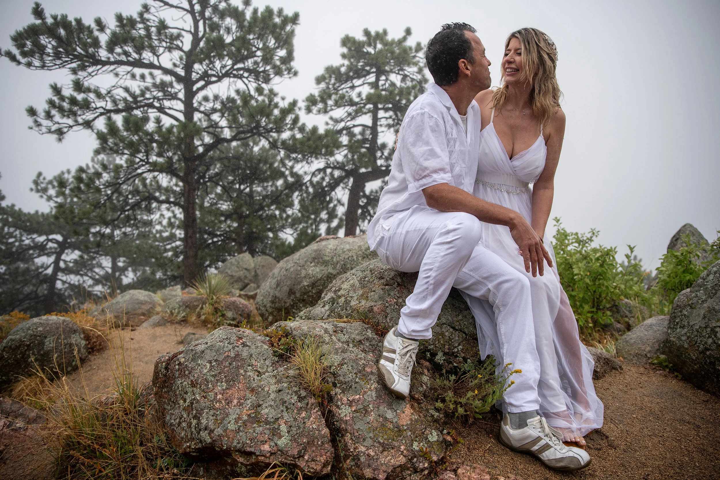 A man and woman dressed in white sitting on rocks outdoors, smiling and looking at each other with trees and fog in the background.