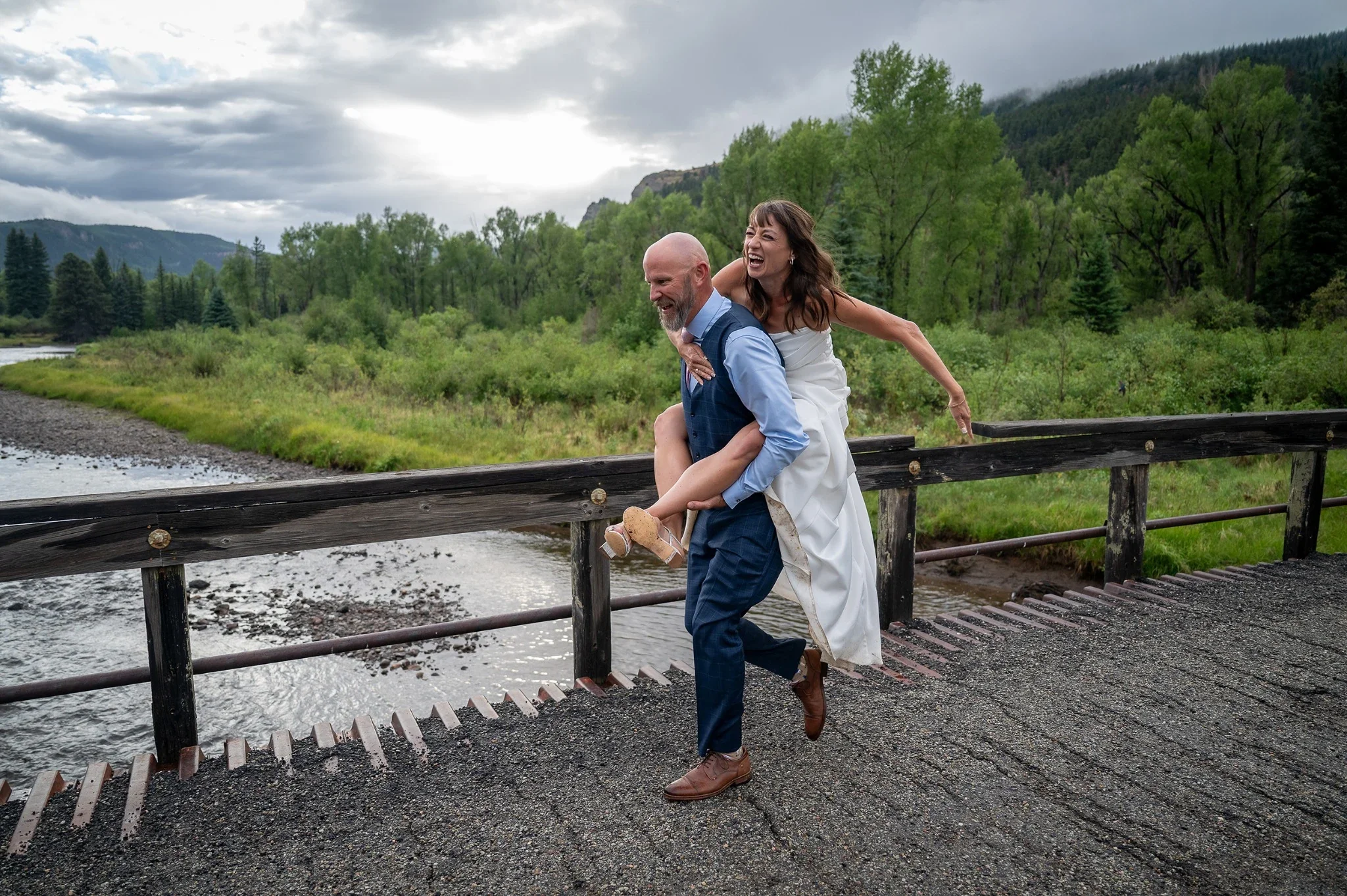 Giddy up! Bride and Groom having fun running across an old bridge on the Conejos River during a summer intimate wedding near Horca, Colorado. Ryan Kost Photography.