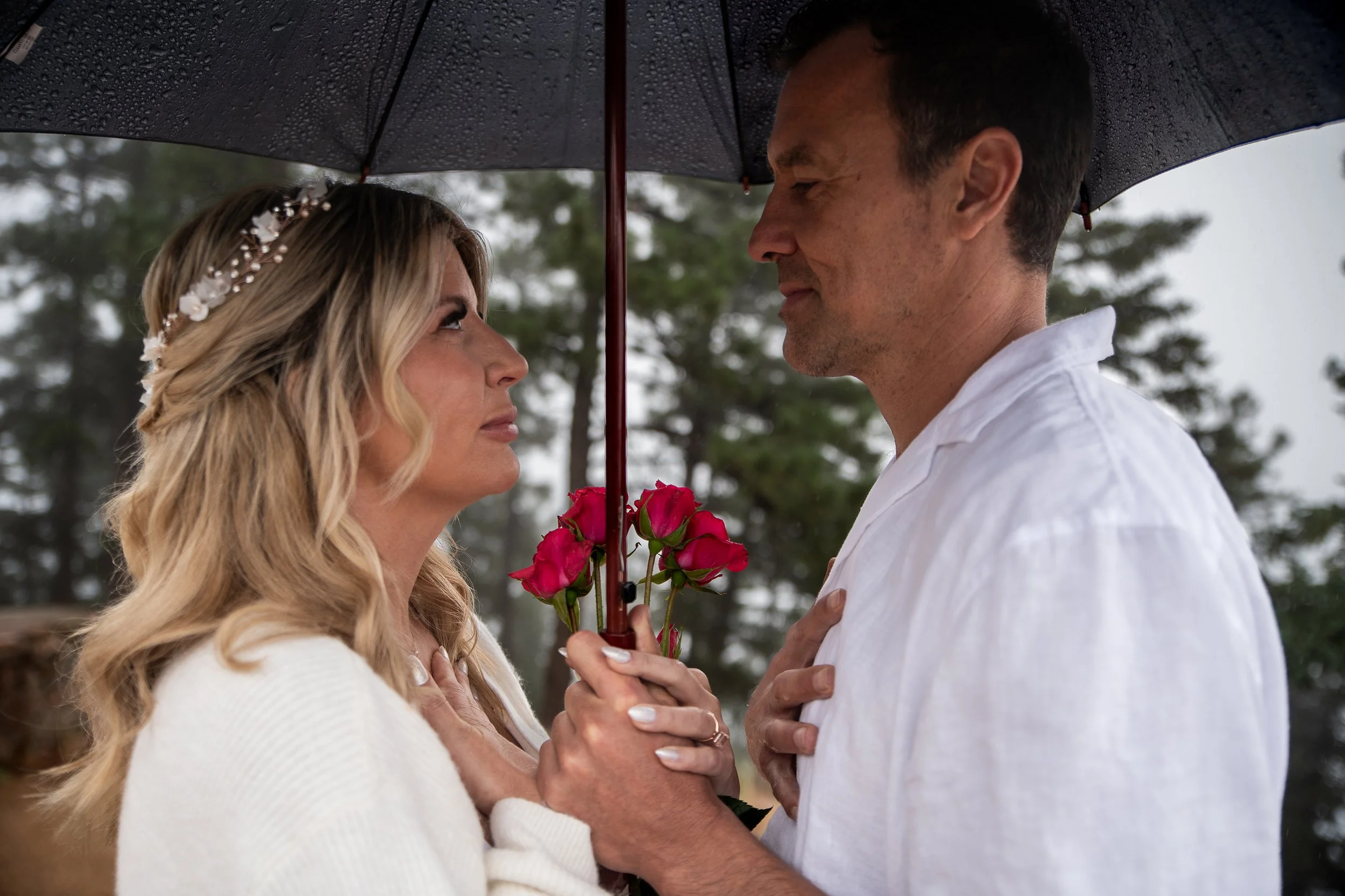 A couple stands close together under a black umbrella on a rainy day, with the woman holding a small bouquet of pink roses, both touching their hearts and gazing into each other's eyes.