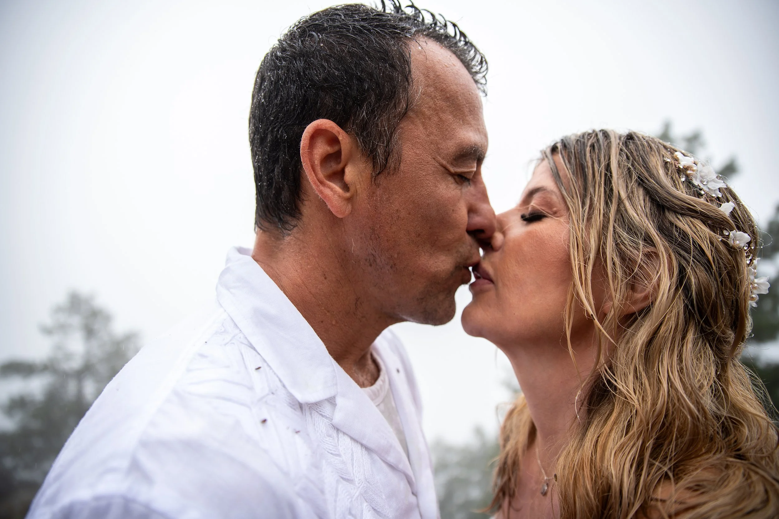 A man and woman are kissing outdoors, with trees and an overcast sky in the background.